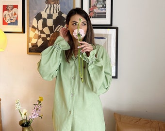May include: A woman wearing a light green, long-sleeved pajama top with a button-down front. She is holding a pink flower in front of her face.