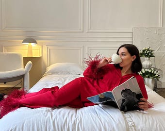 May include: A person wearing a red pajama set with feather trim on the sleeves and pant legs, holding a white teacup and reading a magazine. The person is lying on a white bed with a white pillow and comforter.