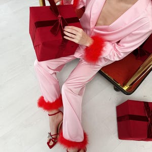 May include: Pink satin pajama set with red feather trim on the cuffs and ankles. The outfit is paired with red heeled sandals with bow details. A red gift box with a velvet ribbon is held in the lap, and another is on the floor.