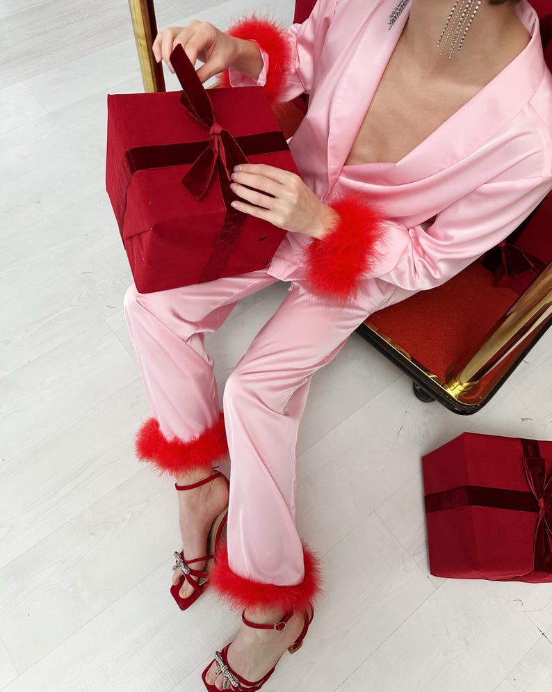 May include: Pink satin pajama set with red feather trim on the cuffs and ankles. The outfit is paired with red heeled sandals with bow details. A red gift box with a velvet ribbon is held in the lap, and another is on the floor.