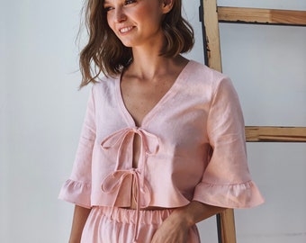 May include: A light pink cropped top with a V-neckline, three-quarter length ruffled sleeves, and two front tie closures, paired with matching high-waisted shorts. The garment features a textured fabric finish and a simple, clean design.