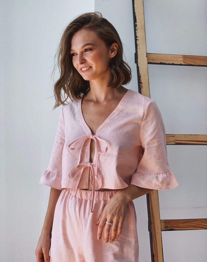 May include: A pink linen crop top with a tie front and ruffled sleeves. The top is paired with matching pink linen shorts.