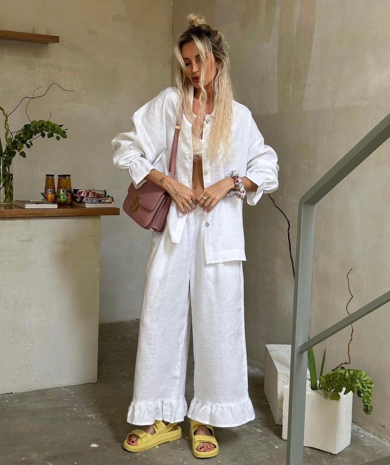 May include: A white linen shirt and pants set with a ruffled hem. The shirt has a relaxed fit and long sleeves. The model is wearing a pink shoulder bag.