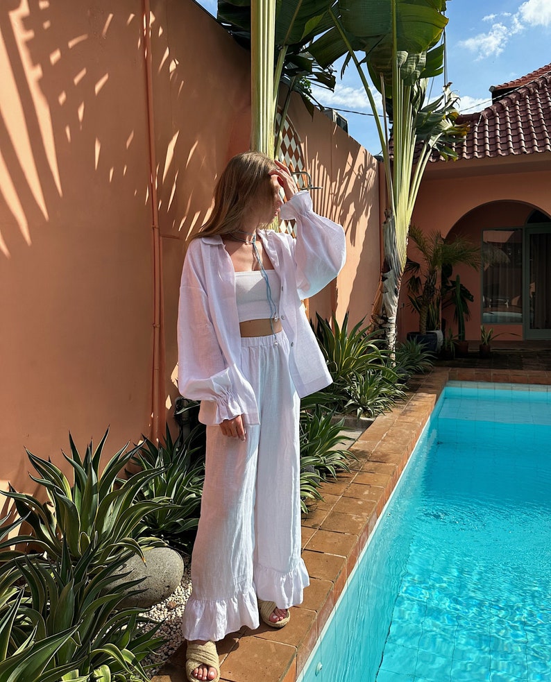 May include: A woman wearing a white linen shirt and pants with ruffled hems. She is standing by a swimming pool with a palm tree in the background.