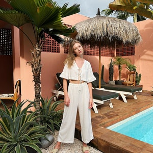 May include: A white linen crop top with a tie front and short sleeves paired with matching wide-leg pants. The model is standing by a pool in a tropical setting.