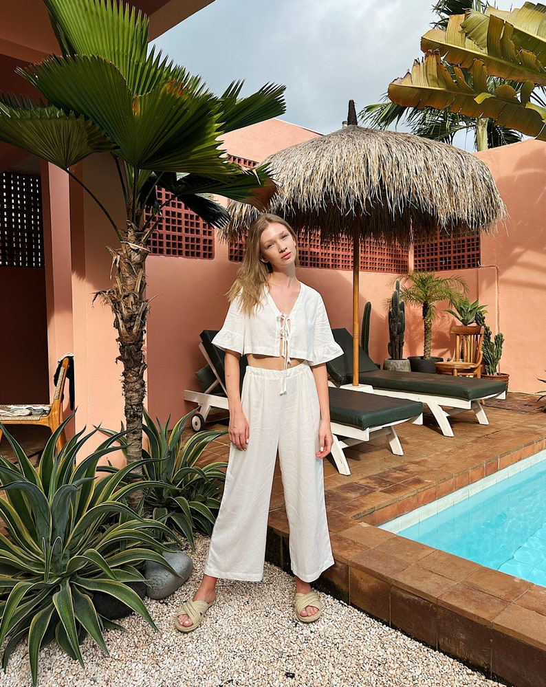 May include: A white linen crop top with a tie front and short sleeves paired with matching wide-leg pants. The model is standing by a pool in a tropical setting.