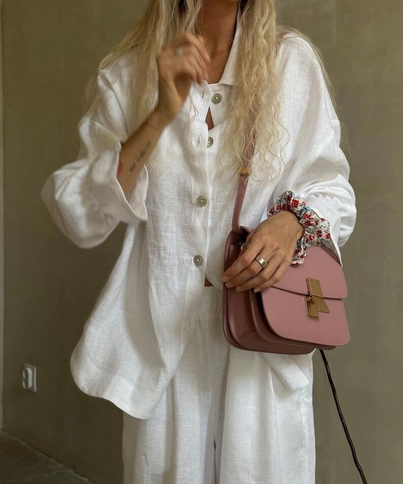 May include: A white linen shirt with a button-down collar and long sleeves. The shirt is worn with matching white linen pants. A pink leather shoulder bag with a gold clasp is worn over the shirt. The bag is hanging from the shoulder and is held in the hand.