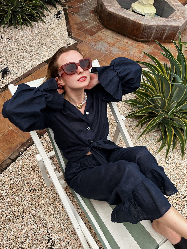 May include: A woman wearing a navy blue linen pantsuit with a ruffled hem and a knotted top. She is wearing brown sunglasses and a pearl and gold necklace. She is sitting on a white lounge chair with a green and white striped cushion.