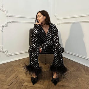 以下が含まれることがあります： 黒と白のドット柄のパジャマセット。パンツには羽のトリムが付いています。黒い蝶ネクタイのヒールと合わせています。モデルは黒い椅子に座っています。