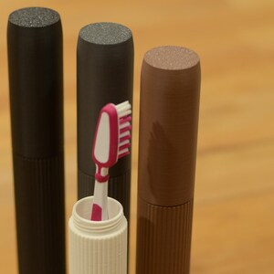 Op de afbeelding: Drie cilindrische reistassen voor tandenborstels. Twee zijn zwart en één is bruin. Eén tas heeft een witte tandenborstel met roze borstels erin.
