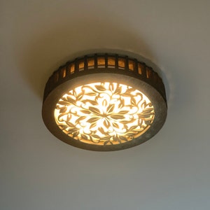 Könnte beinhalten: Eine runde, hölzerne Deckenleuchte mit einem ausgeschnittenen Blumendesign. Das Licht ist eingeschaltet und beleuchtet das Design.