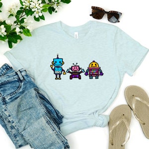 Könnte beinhalten: Ein hellblaues T-Shirt mit einem Motiv von drei Robotern. Die Roboter sind blau, rosa und gelb mit schwarzen Umrissen. Die Roboter lächeln und haben Antennen.