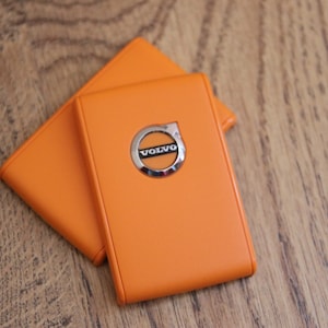 以下が含まれることがあります： オレンジ色の革製ケースが2つ、重ねて配置されています。上のケースには、中央にシルバーのボルボのエンブレムがあります。ケースは木製の表面に置かれています。