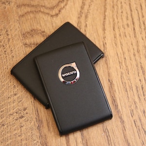 Può includere: Portachiavi in pelle nera con un emblema Volvo argentato. Il portachiavi rettangolare è appoggiato sopra un portafoglio in pelle nera abbinato. Lo sfondo è una superficie di legno marrone chiaro.