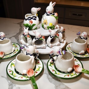 May include: A collection of white ceramic Easter-themed tableware. Includes bunny-shaped egg cups with matching spoons, bunny napkin rings, and decorative serving pieces. The pieces feature colourful floral and checkered accents.