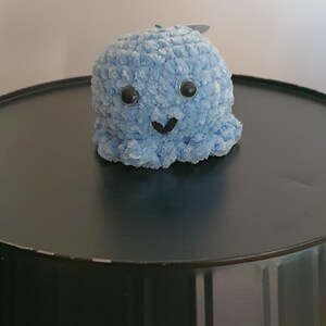 Peut inclure: Une pieuvre au crochet bleu clair faite à la main, avec des yeux globuleux et un sourire cousu. Le jouet en peluche est de forme ronde et douce, idéal pour une chambre d'enfant ou comme cadeau unique. La pieuvre est posée sur une surface sombre.