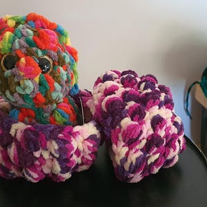 May include: A vibrant, handmade crochet octopus with large, black eyes sits in a matching basket. Beside it is a smaller, purple and white crochet item. The octopus is made of various colours, including red, green, and blue.