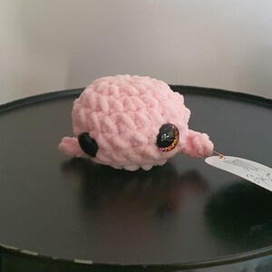 May include: A small, pink crocheted octopus toy with large, glittery eyes and small tentacles. The plushie sits on a dark surface, with a tag attached. The toy is handmade and likely intended for children.
