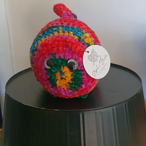Könnte beinhalten: Ein farbenfrohes, handgehäkeltes Raupenspielzeug. Es hat eine Mischung aus roter, rosa, blauer und grüner Wolle mit einem gelb-grünen Zentrum. Das Spielzeug hat große Wackelaugen und ein Etikett mit der Aufschrift "Steph's Studio".