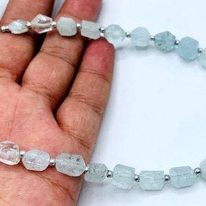 Pode incluir: Um colar com contas facetadas de água-marinha em tons de azul claro e transparente. As contas são enfiadas numa corrente de prata, criando uma joia delicada e elegante. O colar é exibido sobre um fundo branco.
