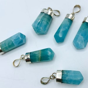 May include: Six blue-green crystal pendants with silver-colored metal caps and loops. The crystals have a faceted, geometric shape. The pendants are arranged on a white surface.