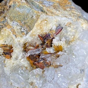 Puede incluir: Primer plano de un espécimen mineral con grupos de cristales naranjas, marrones y rojizos incrustados en una matriz de cuarzo blanco y translúcido. El mineral tiene una estructura cristalina.