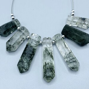 May include: A strand of seven clear quartz crystal beads with dark green inclusions. The crystals are faceted and elongated, connected by a thin silver wire and small silver beads. The crystals have a natural, organic appearance.