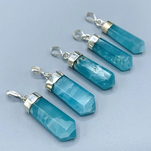 May include: Five blue-green crystal pendants with silver-colored metal caps and loops. The pendants are hexagonal in shape and vary slightly in color and texture. The background is a solid light gray.