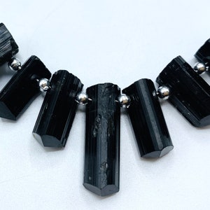 Könnte beinhalten: Eine Halskette mit schwarzen Turmalin-Edelsteinperlen. Die zylindrischen, polierten Steine sind auf einem silberfarbenen Draht aufgereiht, getrennt durch kleine, runde, silberfarbene Perlen. Die Edelsteine haben eine glänzende, reflektierende Oberfläche.