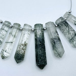 May include: A necklace featuring seven clear quartz crystal pendants with black inclusions. The crystals are faceted and suspended on a thin silver-colored chain with small silver beads. The crystals are approximately 1.5 inches long.