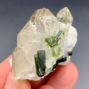 May include: A cluster of quartz crystals with green tourmaline inclusions. The clear quartz points are accented by dark green, elongated tourmaline crystals and smaller green clusters. The base features white and black mineral deposits.