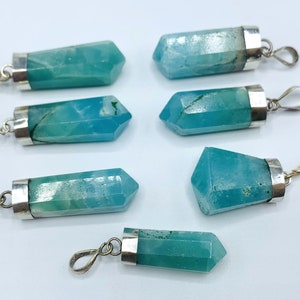 May include: Seven blue-green crystal pendants with silver-colored metal caps and loops. The crystals are hexagonal and faceted, with a polished appearance. The pendants are arranged on a white surface.