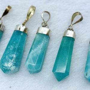 May include: Five aquamarine crystal pendants with silver-colored metal caps and loops. The pendants are a light blue-green color and have a faceted, elongated shape. The pendants are displayed on a white surface.