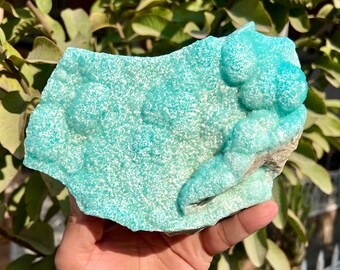 Op de afbeelding: Een groot, onregelmatig stuk turkooiskleurig mineraal met een textuur, bubbelachtig oppervlak. Het mineraal heeft een ruwe, natuurlijke rand en een lichtgekleurde basis. De algemene uitstraling is die van een ruwe, ongepolijste steen, waarschijnlijk een exemplaar voor verzamelaars of decoratief gebruik.