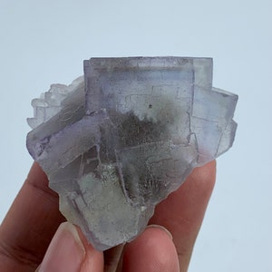 May include: A close-up of a fluorite crystal cluster. The crystal has a pale purple hue with some areas appearing translucent. The crystal structure is visible, with distinct cubic formations. The background is a plain white.