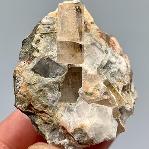 May include: A raw mineral specimen featuring a light brown crystal formation protruding from a matrix of gray, white, and tan rock. The crystal has a rectangular shape and a translucent appearance. The rock has a rough, textured surface.