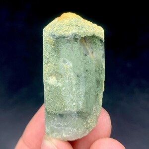 Puede incluir: Un cristal de cuarzo verde con una superficie rugosa y texturizada. El cristal tiene un tono verde claro con áreas de cuarzo amarillo y transparente. El cristal se sostiene en la mano de una persona contra un fondo oscuro.