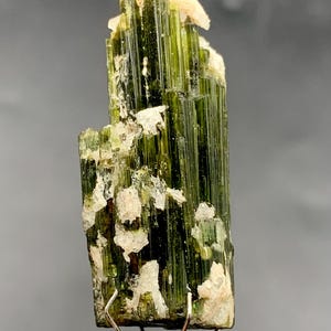 May include: A vibrant green tourmaline crystal specimen with a columnar structure. The crystal has a rich, deep green color with white and tan patches. The mineral is mounted on a silver metal stand, showcasing its natural beauty.