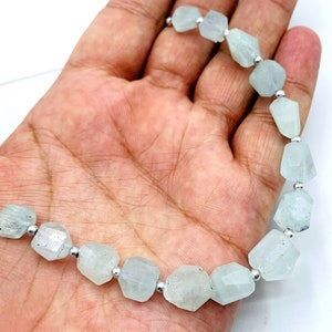 May include: A strand of aquamarine gemstone beads. The beads are faceted, irregular shapes in a pale blue-green color. Each bead is separated by a small, silver-colored spacer. The strand is held against a white background.