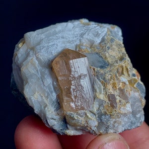 May include: A natural mineral specimen featuring a large, translucent, light brown crystal with a prismatic shape. The crystal is embedded in a matrix of white and gray rock, with hints of tan and other minerals. The overall composition is rough and textured.