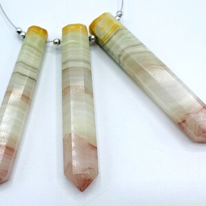 May include: Three faceted, elongated crystal pendants. The crystals are primarily white with bands of light brown, and a reddish-pink tip. The top of each crystal is a yellow-orange color. The crystals are strung on a thin, silver-colored wire with small silver beads.