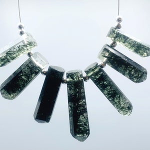 Può includere: Una collana con sette perline di cristallo allungate e sfaccettate. Le perline sono trasparenti con inclusioni verde scuro, infilate su un sottile filo d'argento con piccole perline d'argento tra ogni cristallo. I cristalli sono disposti a ventaglio.