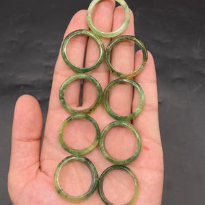 Puede incluir: Una colección de ocho anillos de jade en varios tonos de verde. Los anillos son circulares, con una superficie lisa y pulida. Los anillos se muestran en una mano sobre un fondo oscuro. Los anillos son de color verde natural.