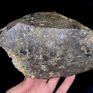 May include: A large, rough-hewn, translucent mineral specimen with a dark gray and gold color palette. The surface has a textured appearance with visible inclusions and a polished edge. The overall shape is irregular, with a flat, cut face.