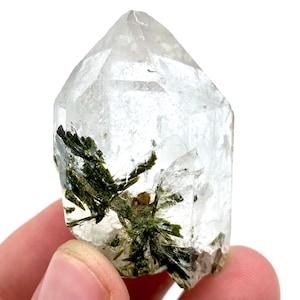 Puede incluir: Un cristal de cuarzo transparente con inclusiones verdes. El cristal es transparente con una parte superior puntiaguda y una base rugosa. Las inclusiones verdes son en forma de aguja y están agrupadas en todo el cristal.