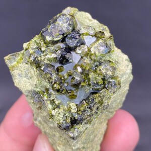 Puede incluir: Primer plano de un espécimen mineral en bruto. La piedra presenta grupos de cristales verdes oscuros y negros sobre una matriz verde claro. Los cristales tienen un aspecto reflectante y vítreo, y la textura general es rugosa y natural.