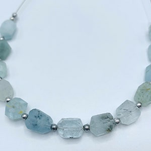 Pode incluir: Um colar de contas de pedra preciosa água-marinha. As contas são uma mistura de azul pálido e verde mar, com uma forma irregular facetada. Cada conta é separada por uma pequena conta prateada. O fio é fino e transparente.