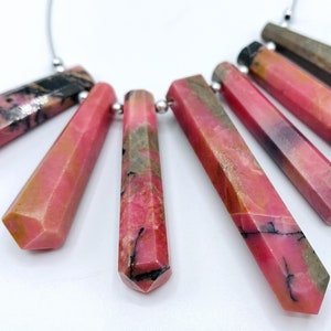 Pode incluir: Uma coleção de pingentes de pedras preciosas rosa e pretas, cada um com uma forma facetada e alongada. As pedras são enfiadas em uma corrente prateada, e os pingentes exibem uma mistura de tons rosa, preto e castanho. As pedras preciosas estão dispostas em forma de leque.