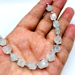 Pode incluir: Um colar de contas de pedras preciosas azul claro e transparentes com espaçadores prateados. As contas têm formas irregulares e facetadas, enfiadas num cordão fino e transparente. O fundo é branco.