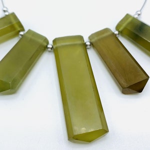 May include: A necklace featuring five olive-green, rectangular-shaped beads with faceted edges. The beads are strung on a silver-colored chain, with a polished finish. The beads have a translucent appearance and a geometric design.